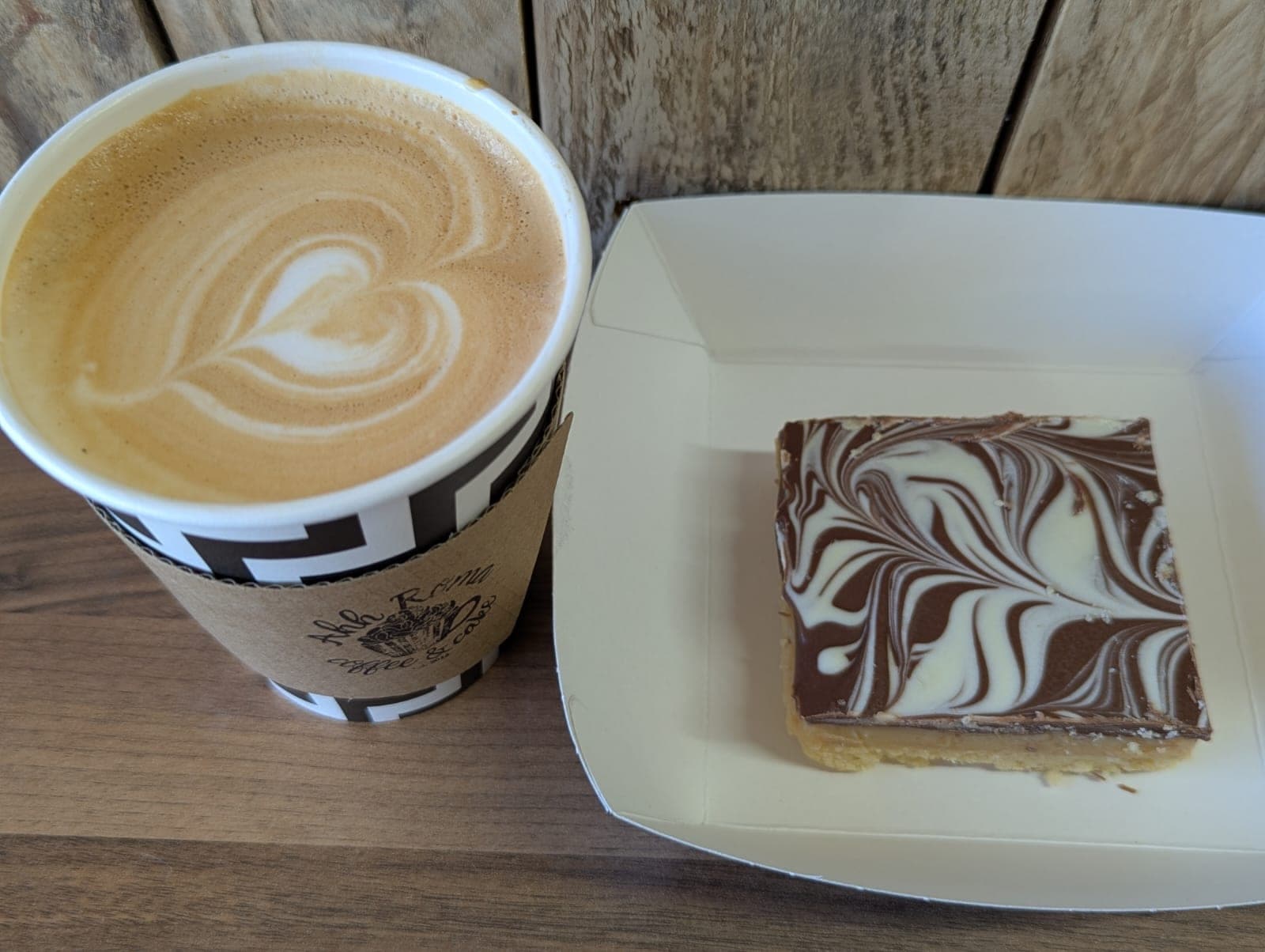 Heart latte art in an Ahh Roma cup beside a slice of marble millionaire's shortbread